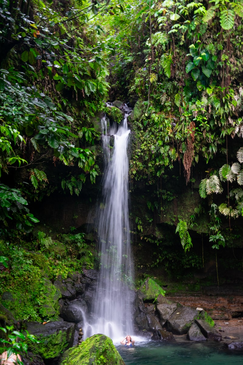 Person standing under the waterfall at Emerald Pool in Dominica surrounded by lush greenery