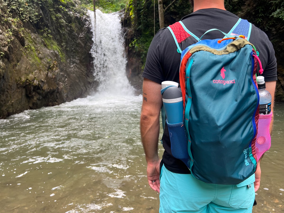 man wearing a Cotopaxi backpack standing in front of a waterfall