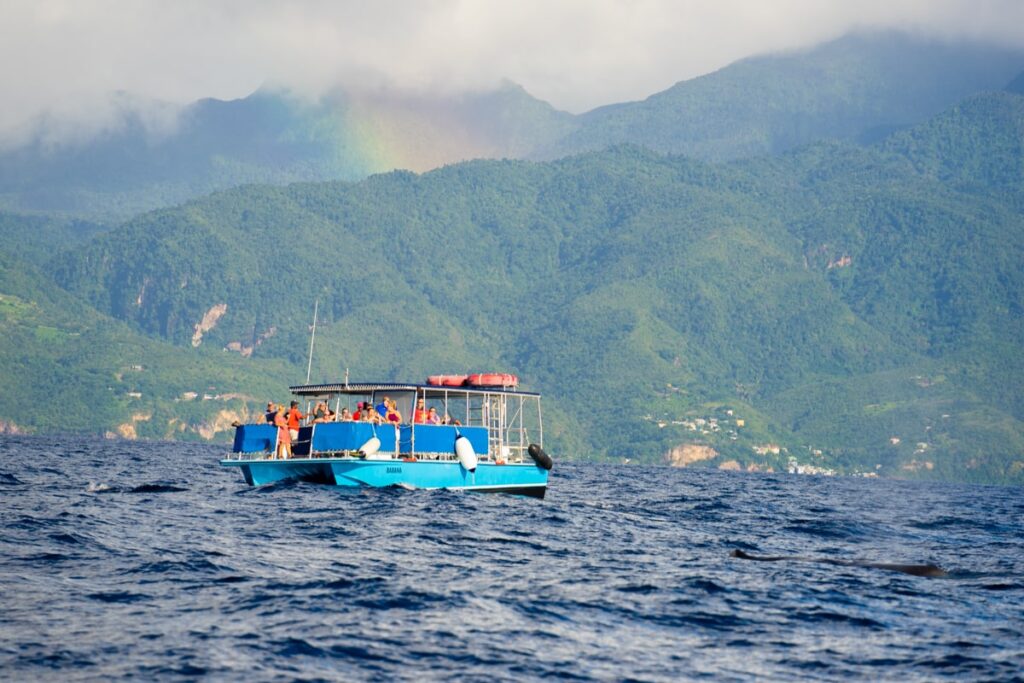 a boat with people floating on the ocean with a rainbow disappearing into the mountains of Dominica behind it