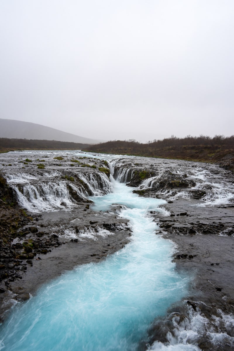 view of the Bruarfoss waterfall from the bridge with bright icy blue water and a cloudy misty sky