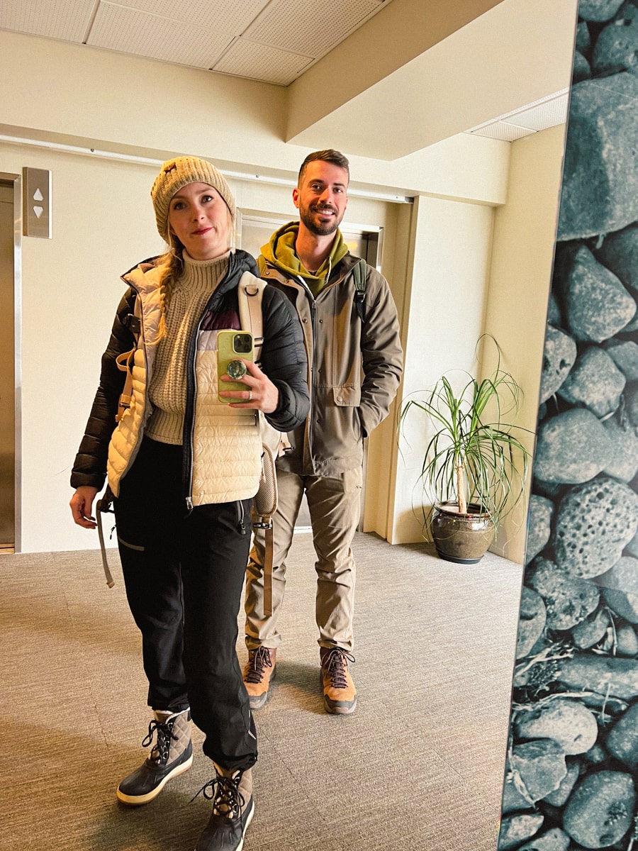 wife and husband taking a photo in a hotel dressed in multiple layers with rain resistant pants, boots, and jackets for the weather in Iceland