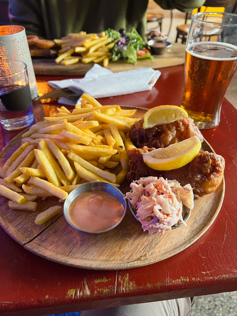 fish and chips meal with coleslaw and a dipping sauce served alongside a beer