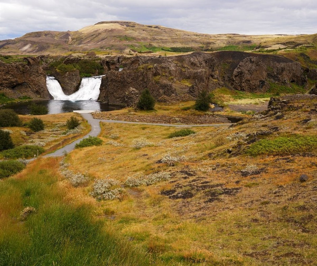 viuew of Hjálparfoss waterfalls joining together and cascading into a pool in Iceland surrounded by green hills 