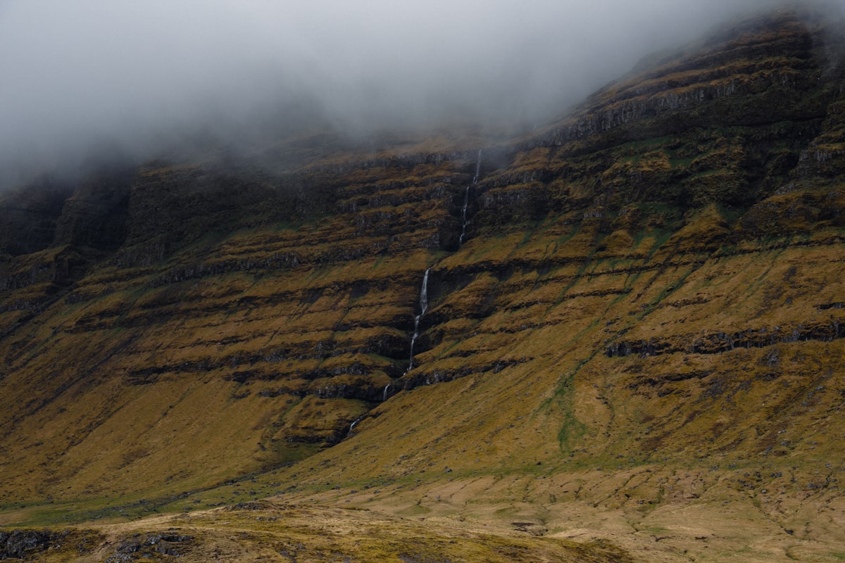 a very tall mountain in iceland with a waterfall streaming down a crevice with a misty cloud at the top