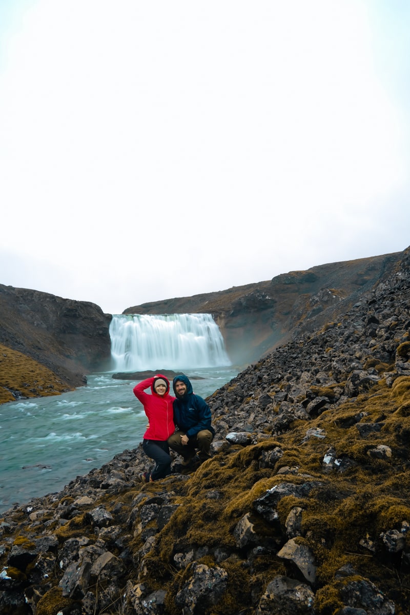 a couple with red and blue jackets crouching down in front of the Porufoss waterfall in Iceland on a cloudy day