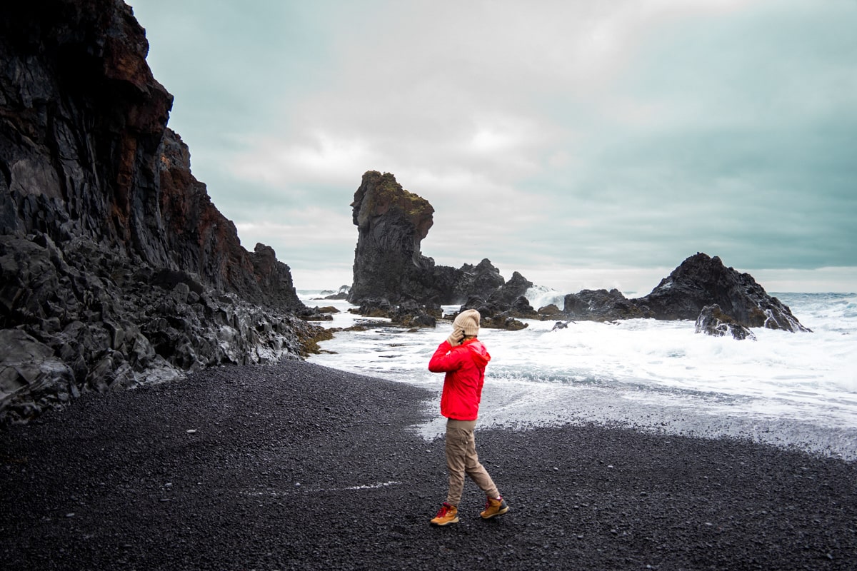 woman wearing a red rain jacket with tan hiking pants, a beanie, and gloves standing on a black sand beach in Iceland