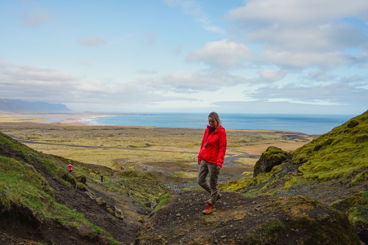 woman in a red jacket standing at the top of a mountain at Reaudfeldsgja Gorge in Snaefellsnes Peninsula iceland overlooking the ocean and landscapes
