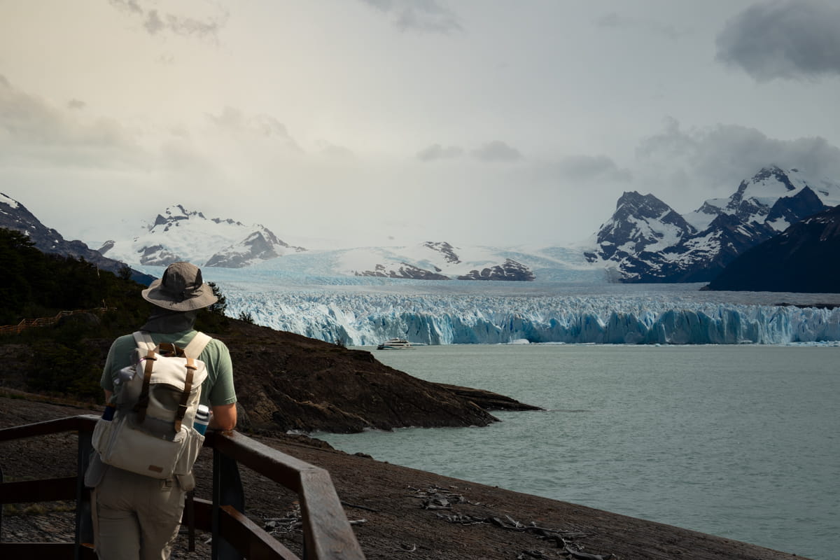 man with backpack and hat standing on a boardwalk overlooking the Perito Moreno glacier in Argentina