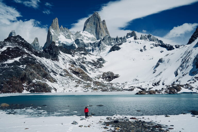 woman in a red jacket standing at the base of Mount Fitz Roy in Patagonia with blue lake