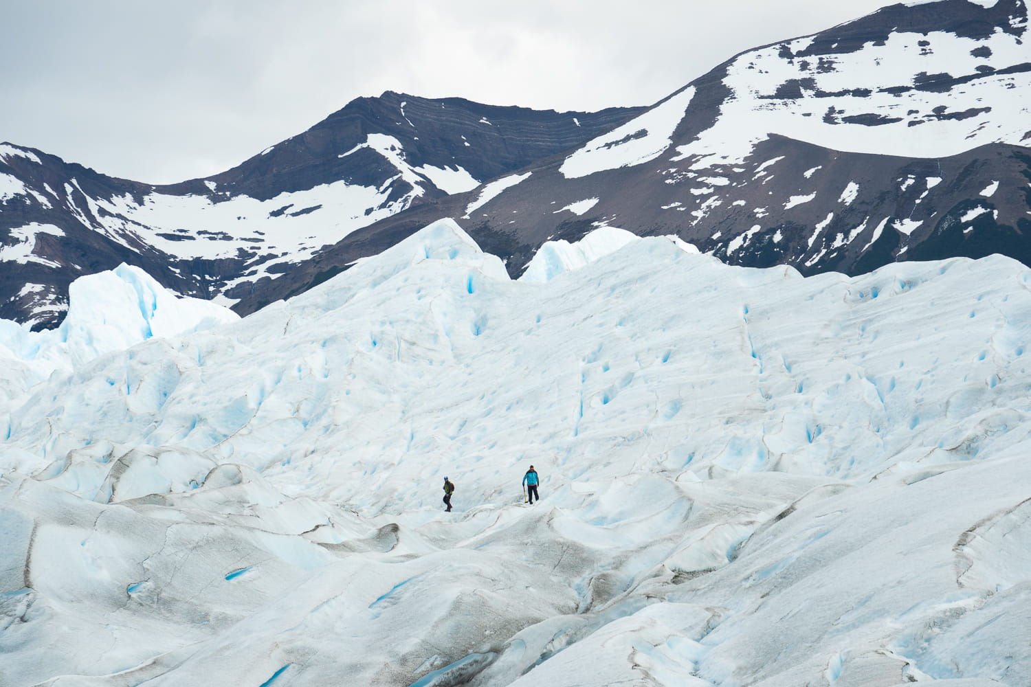 People trekking along the Perito Moreno glacier in the distance with snow-capped mountains in the background