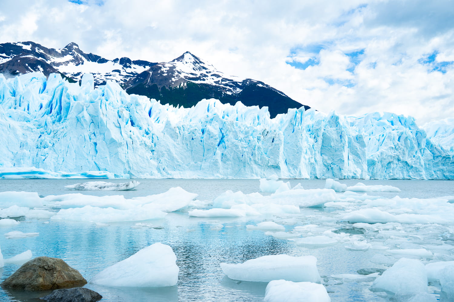 View of the Perito Moreno glacier from water level with floating ice and snow capped mountains in the background